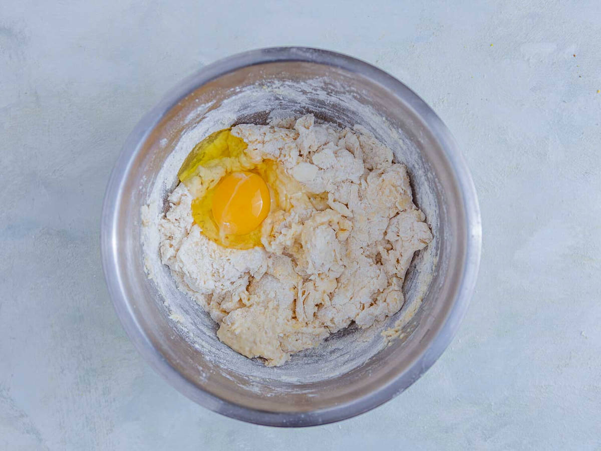 A whole egg cracked onto a shaggy biscuit dough in a stainless steel mixing bowl.