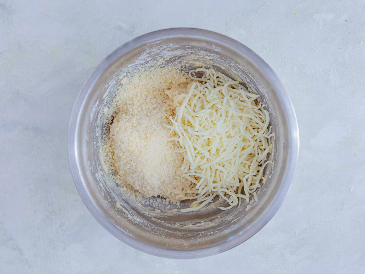 Grated parmesan and shredded mozzarella piled on top of biscuit dough in a mixing bowl, ready to be stirred in.