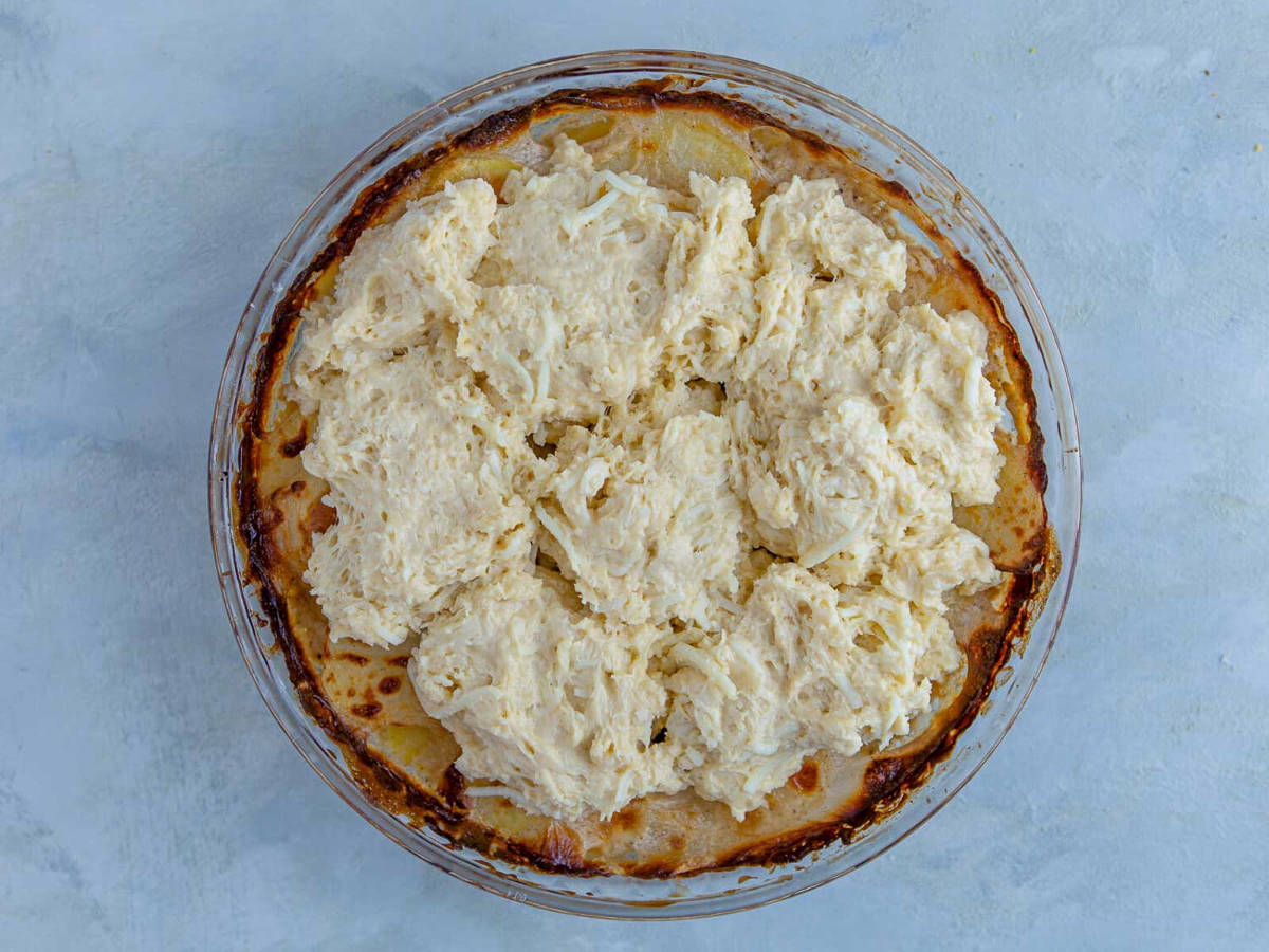 Mounds of cheesy biscuit dough dropped over par-baked potatoes in a glass pie plate, with browned cream around the edges.