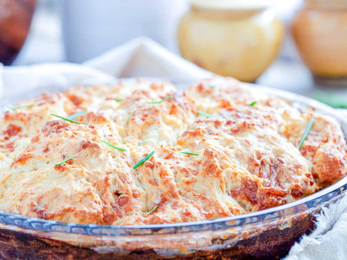 A whole baked potato cobbler in a glass pie plate with a deeply golden, craggy cheese biscuit topping and a few rosemary leaves on top.