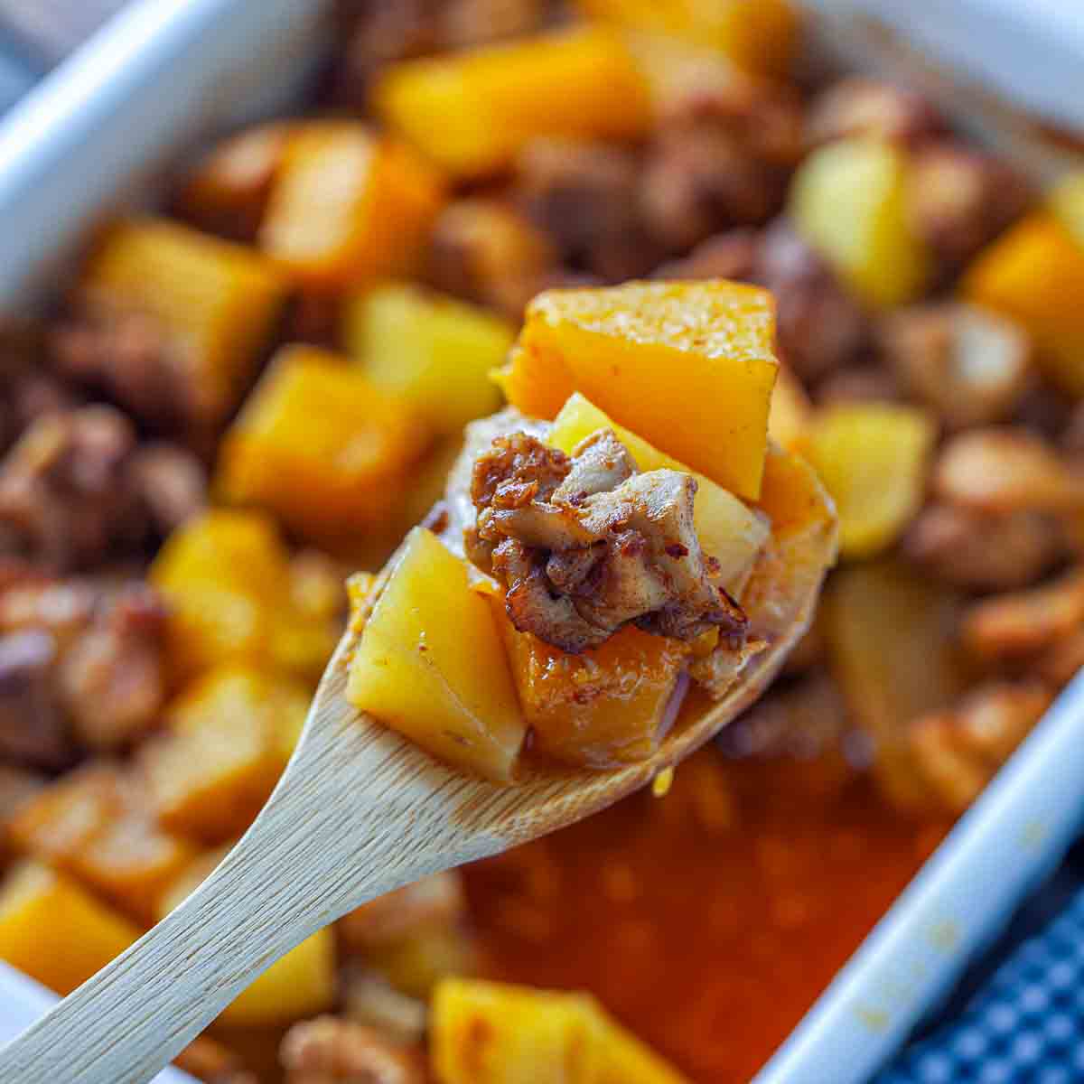 Wooden spoon lifting a bite of chicken, pumpkin, and potato coated in orange-red sauce from the baking dish.
