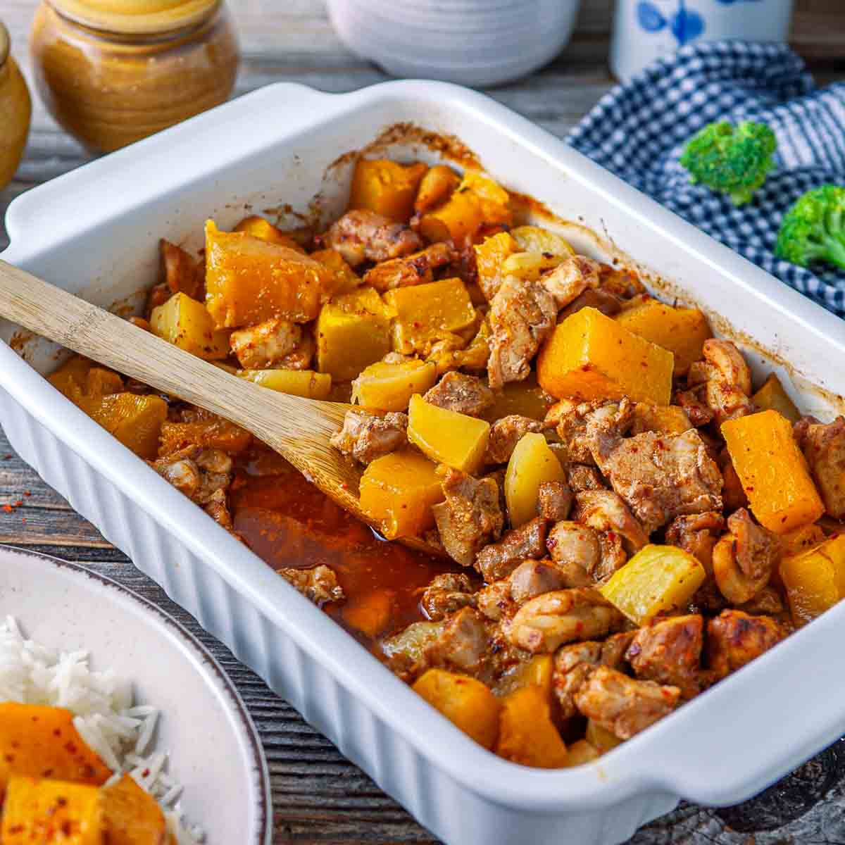 Baking dish of pumpkin chicken casserole with a wooden spoon, next to a plate of rice and pumpkin.