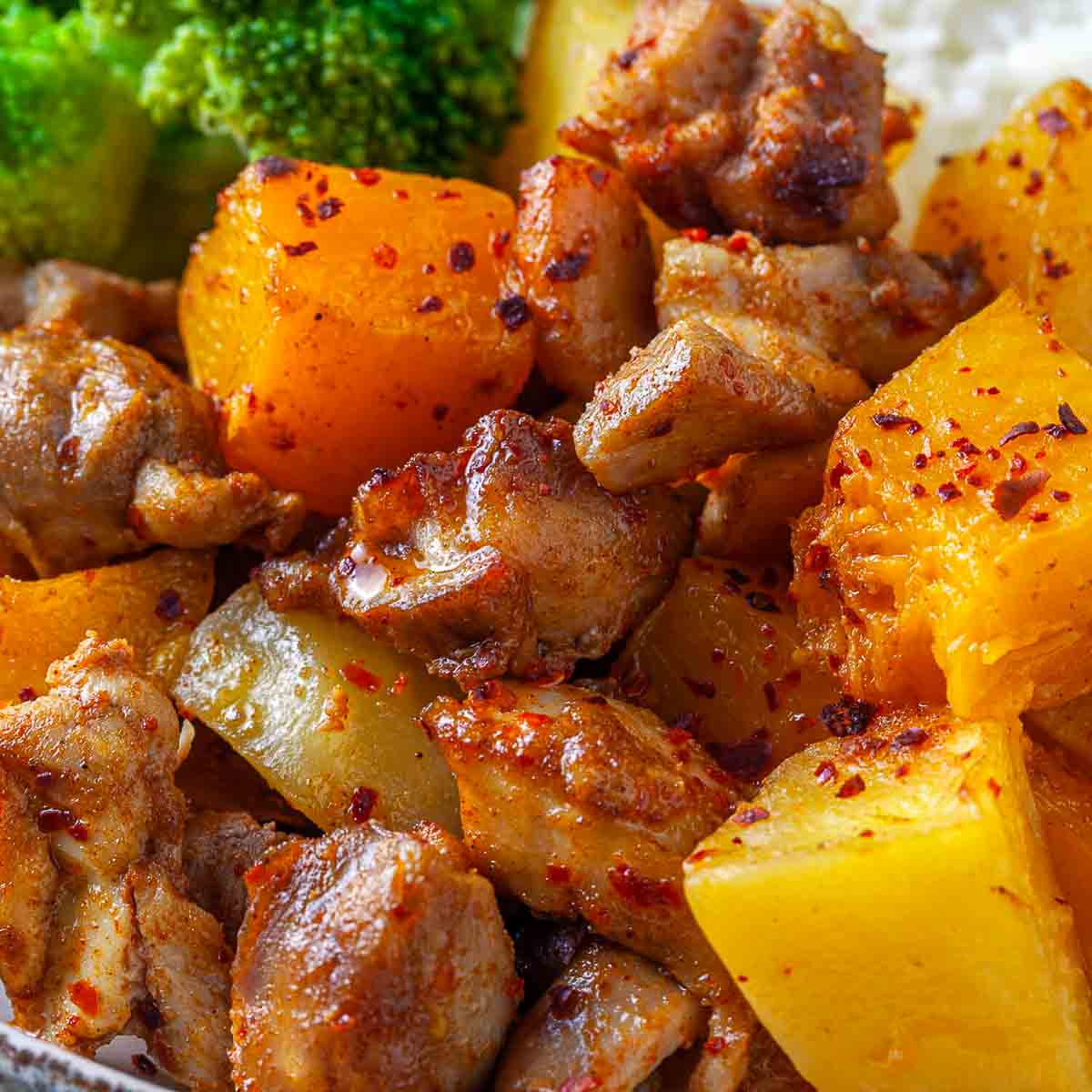 Close-up of glazed chicken, pumpkin, and potato pieces with chili flakes, broccoli visible in the background.