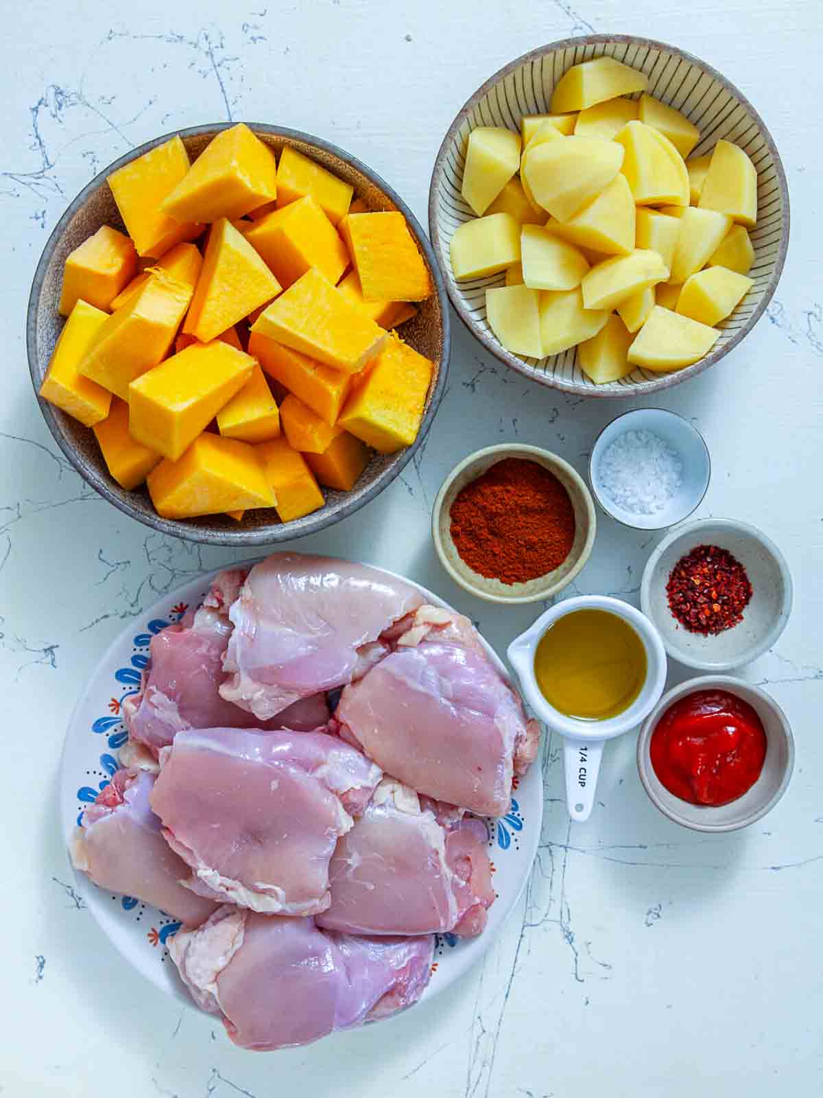 Raw pumpkin and potato cubes, chicken thighs, and spice bowls with paprika, chili flakes, sriracha, and olive oil laid out on a marble counter.