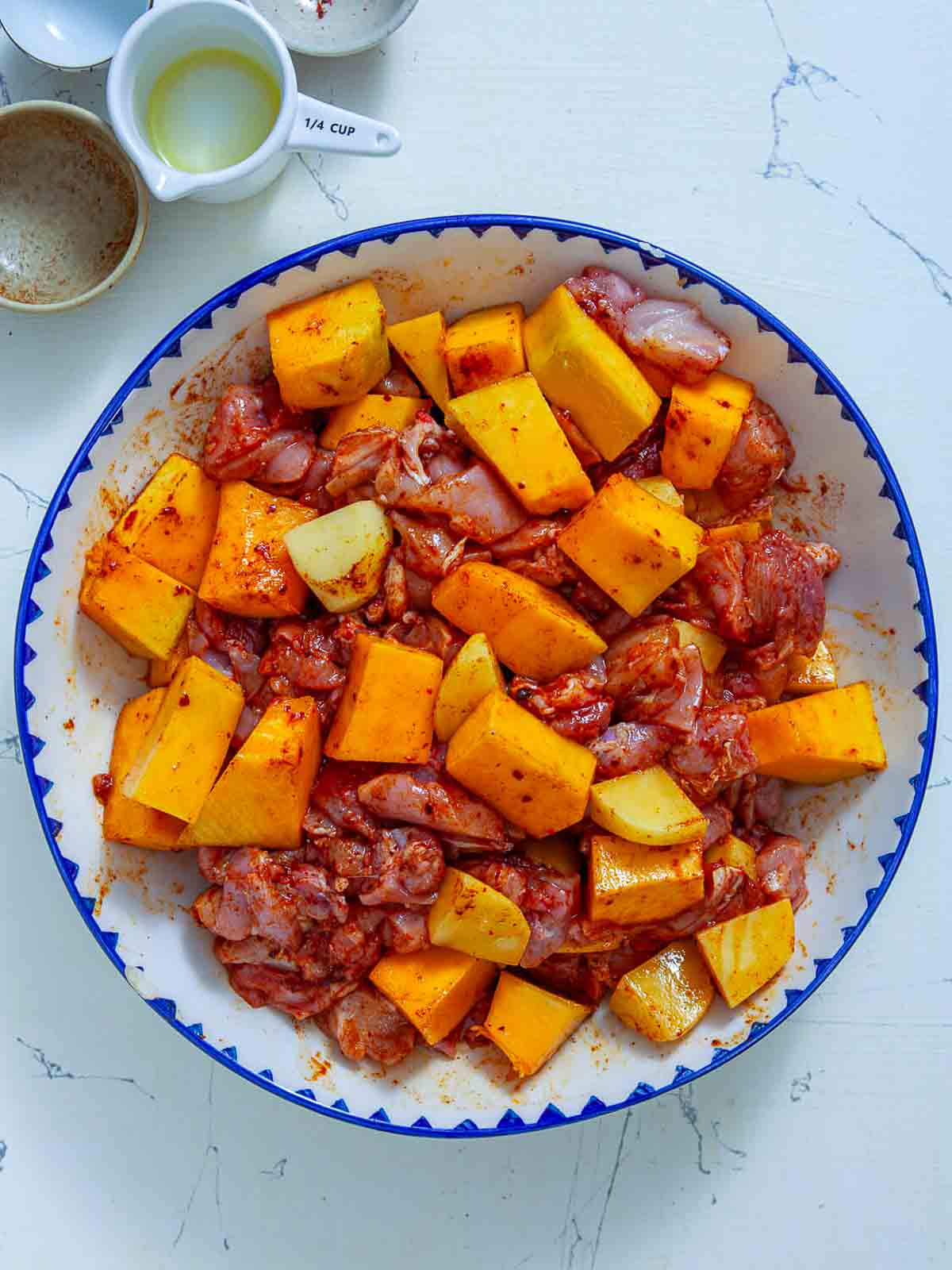 Chicken thighs, pumpkin cubes, and potato cubes tossed in a red sriracha and paprika marinade in a large mixing bowl.