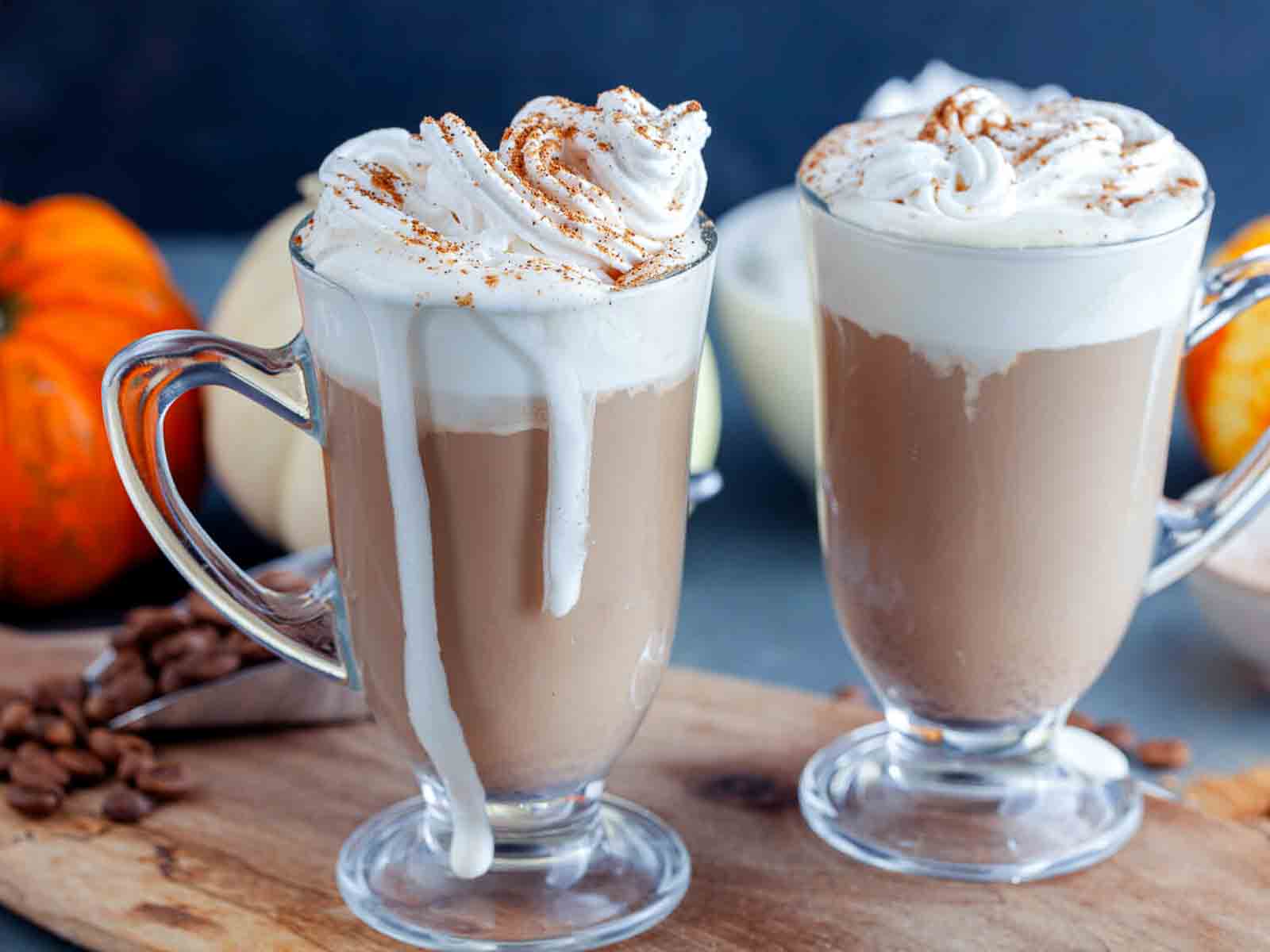 Two footed glass mugs of creamy coffee topped with whipped cream and ground spice on a wooden board scattered with coffee beans.