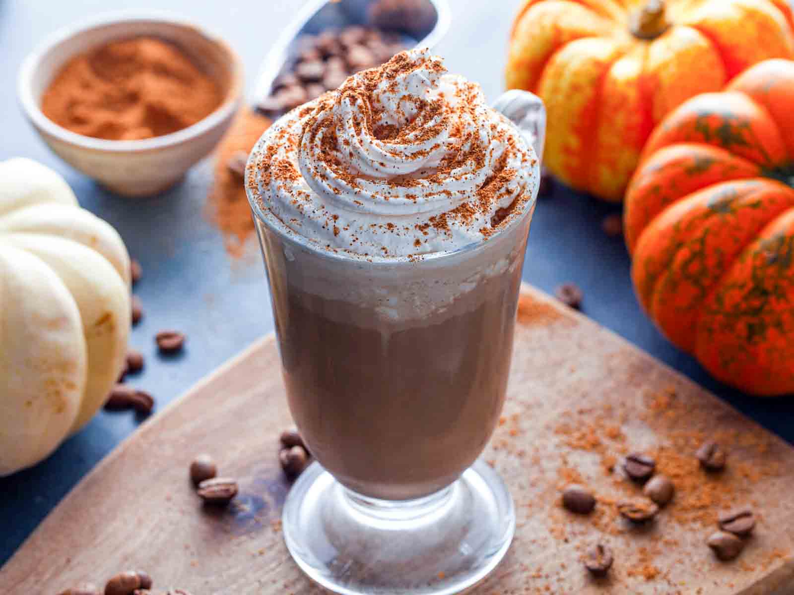  A glass mug of spiced coffee topped with whipped cream and spice, surrounded by white and orange pumpkins, coffee beans, and a bowl of ground cinnamon.