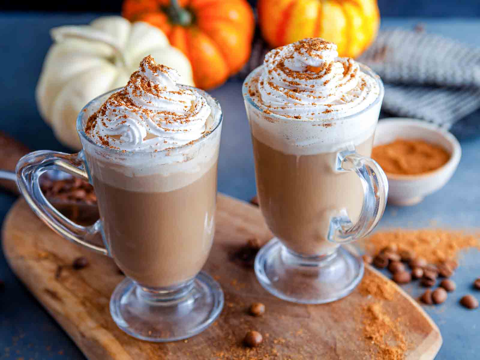 Two glass mugs of creamy coffee with whipped cream overflowing and dripping down the side of the front mug, with pumpkins in the background.