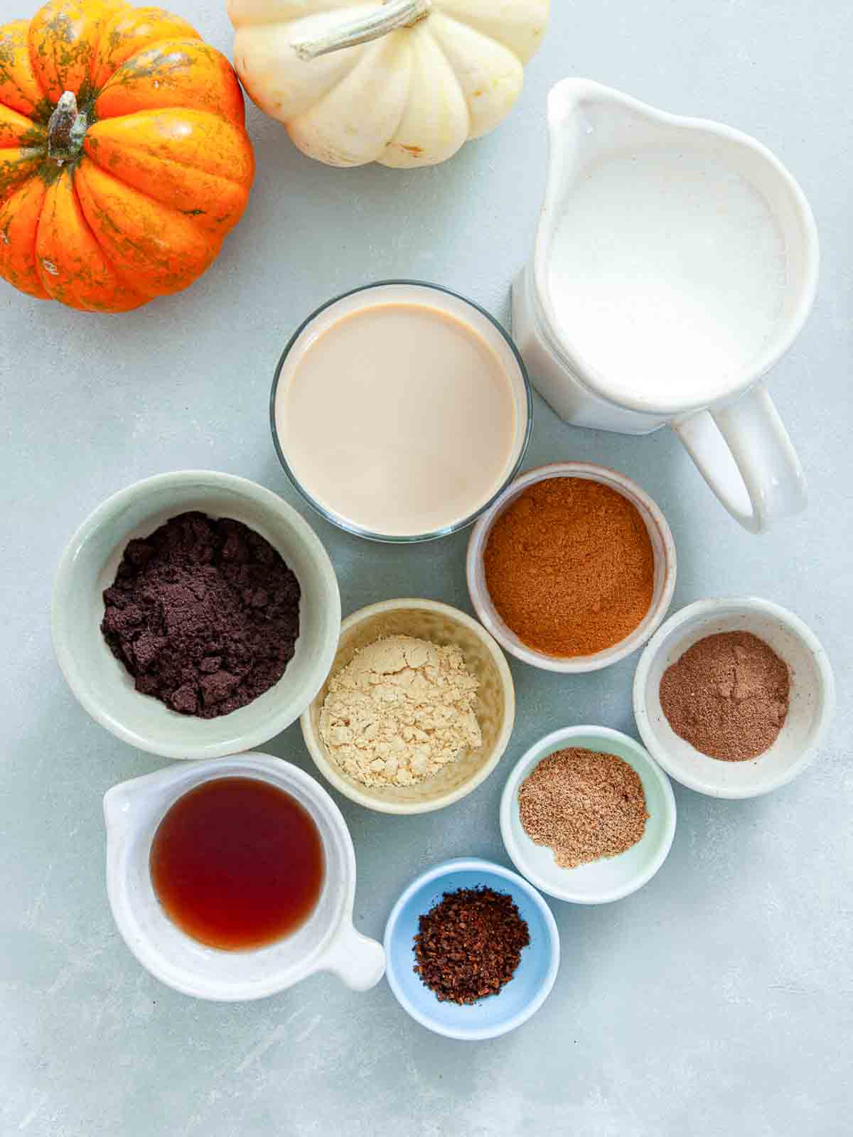 Overhead view of small bowls of ground cinnamon, ginger, nutmeg, allspice, cloves, coffee, and maple syrup beside a glass of Irish cream and a pitcher of cream.