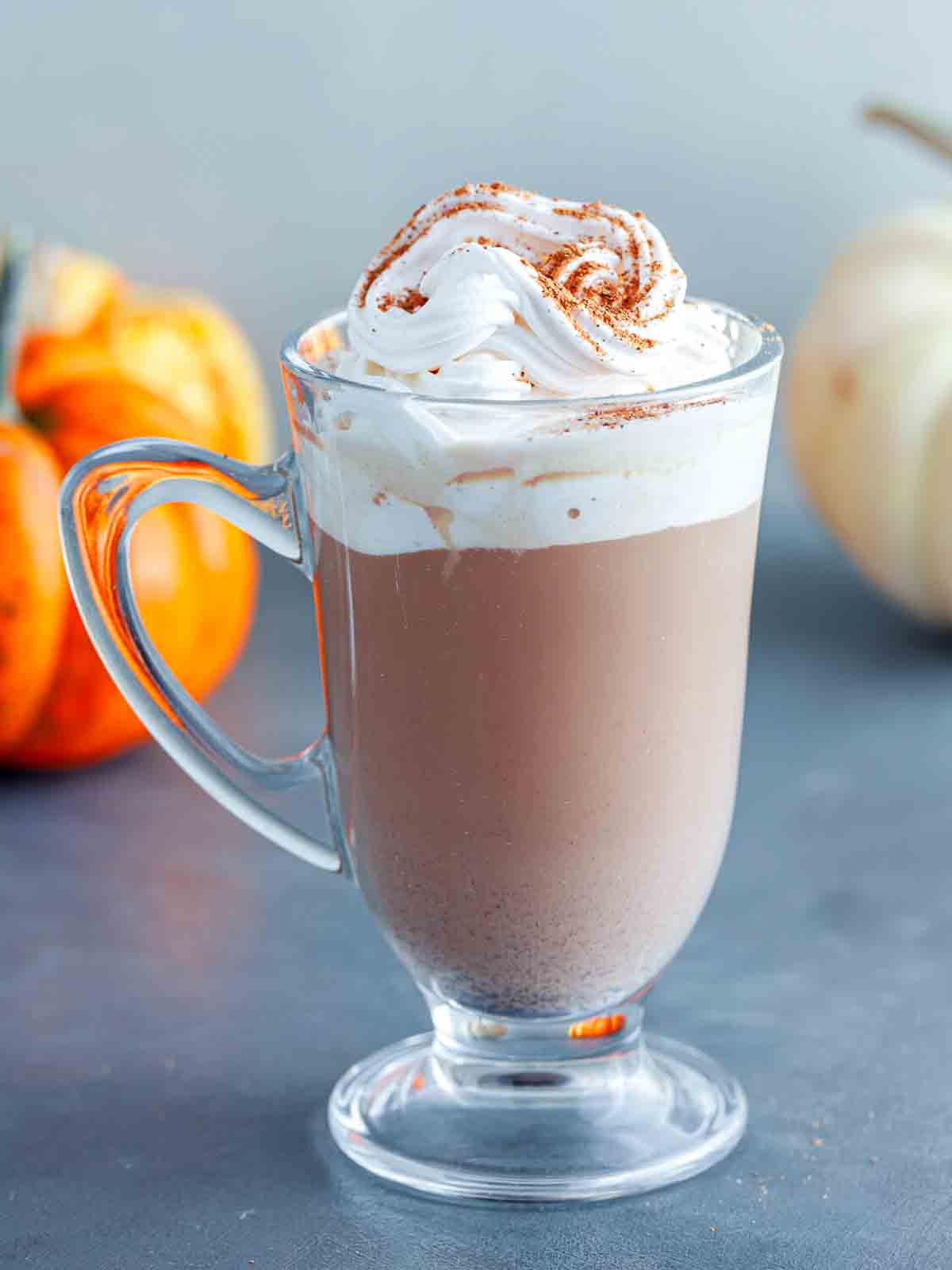 A finished glass mug of spiced coffee with a whipped cream swirl dusted with spice, with pumpkins blurred in the background.
