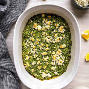 Baked spanakopita dip with golden feta crumbles in a white oval casserole dish, with lemon wedges and a small bowl of crumbled feta beside it.