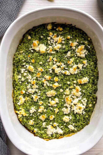Baked spanakopita dip with golden feta crumbles in a white oval casserole dish, with lemon wedges and a small bowl of crumbled feta beside it.