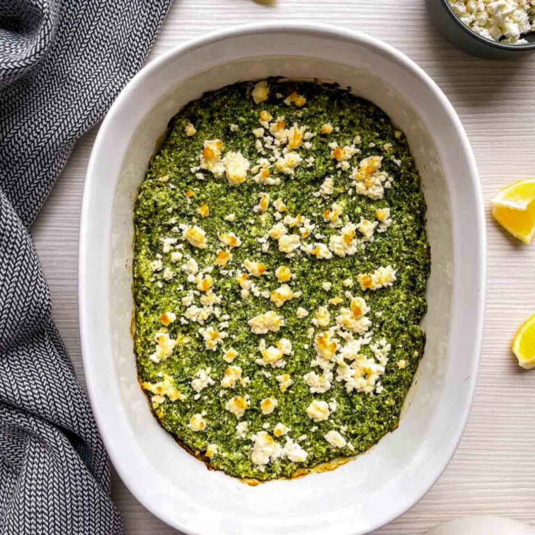 Baked spanakopita dip with golden feta crumbles in a white oval casserole dish, with lemon wedges and a small bowl of crumbled feta beside it.
