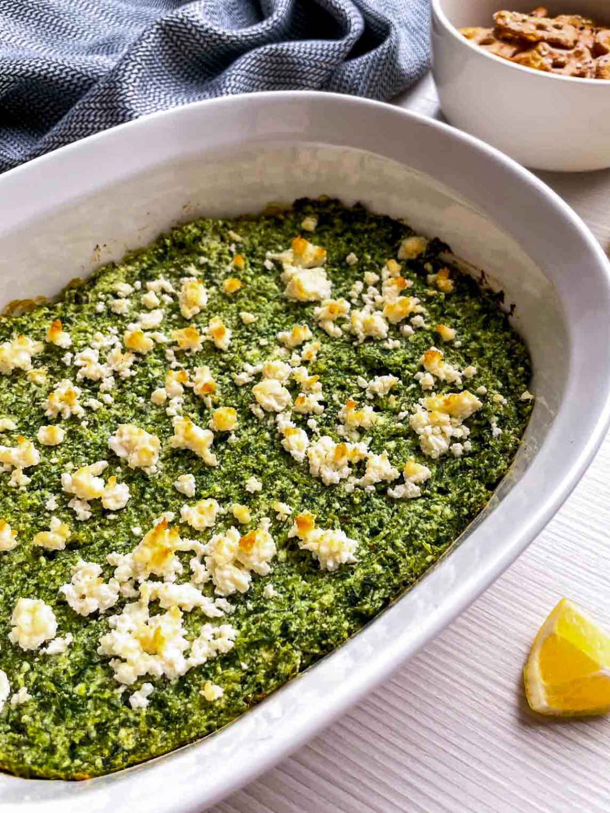 An angled view of a baked spinach and feta dip in a white dish, with a lemon wedge in front and a bowl of pretzel crisps behind.