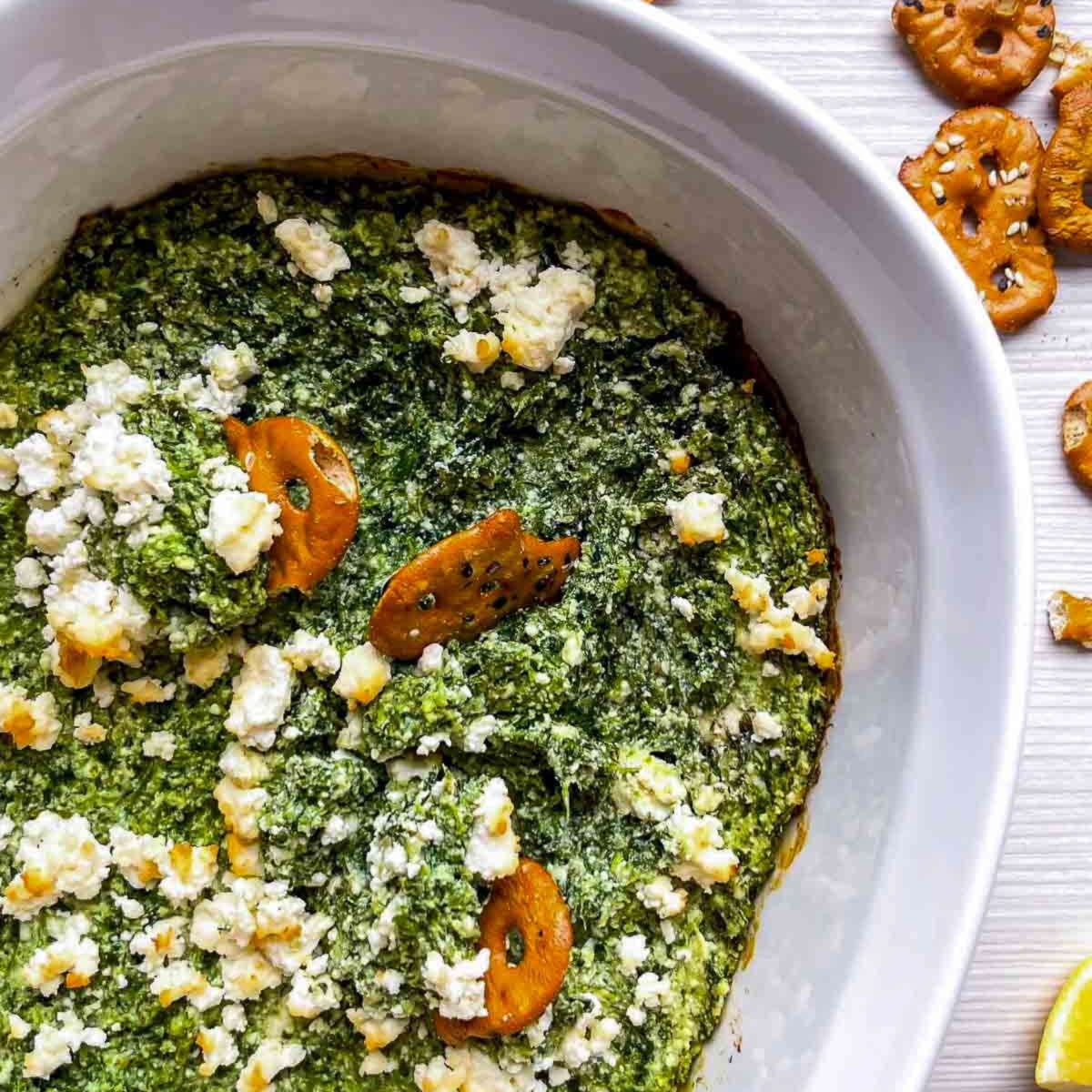 Close-up of a hot green spinach dip topped with browned feta crumbles, with pretzel crisps pressed into the surface.