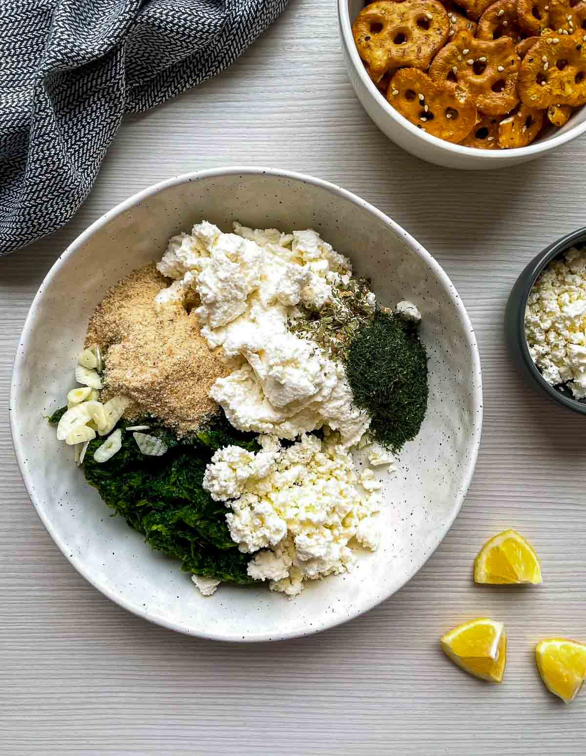 Spinach, ricotta, crumbled feta, sliced garlic, breadcrumbs, dried dill, and oregano piled separately in a speckled white bowl.]