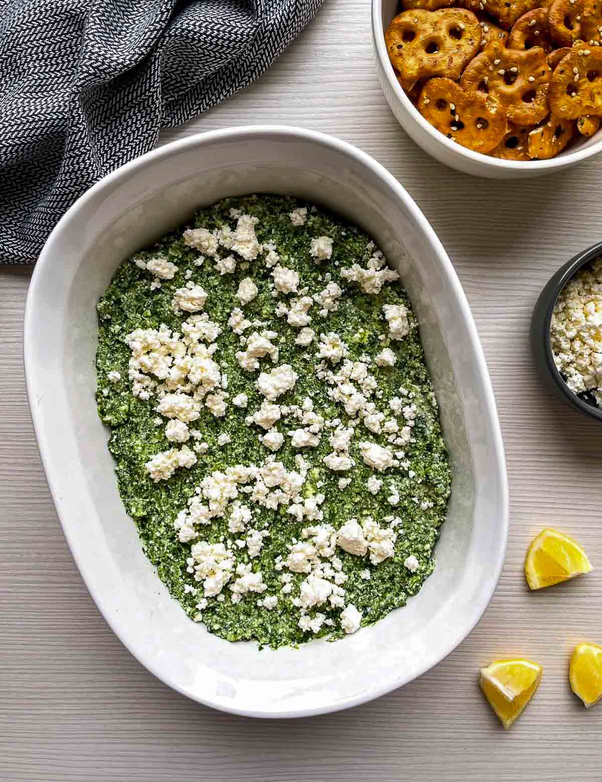 An unbaked spinach mixture spread in a white oval dish and scattered with crumbled white feta.]