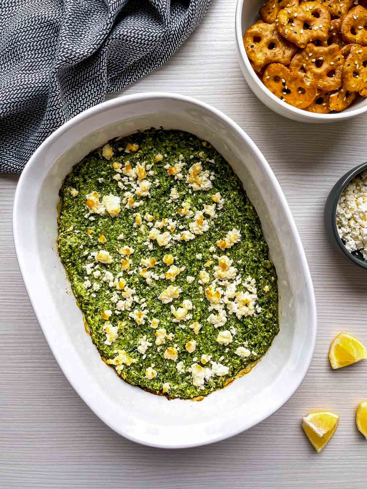A baked spinach dip with golden, toasted feta on top in a white oval dish, next to a bowl of pretzel crisps.